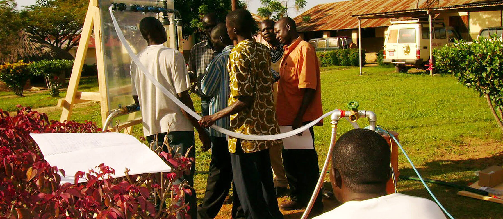 A group of people are standing outside around a water treatment plant, observing and discussing it. A person is sitting in the foreground; buildings, green spaces and a white vehicle can be seen in the background.