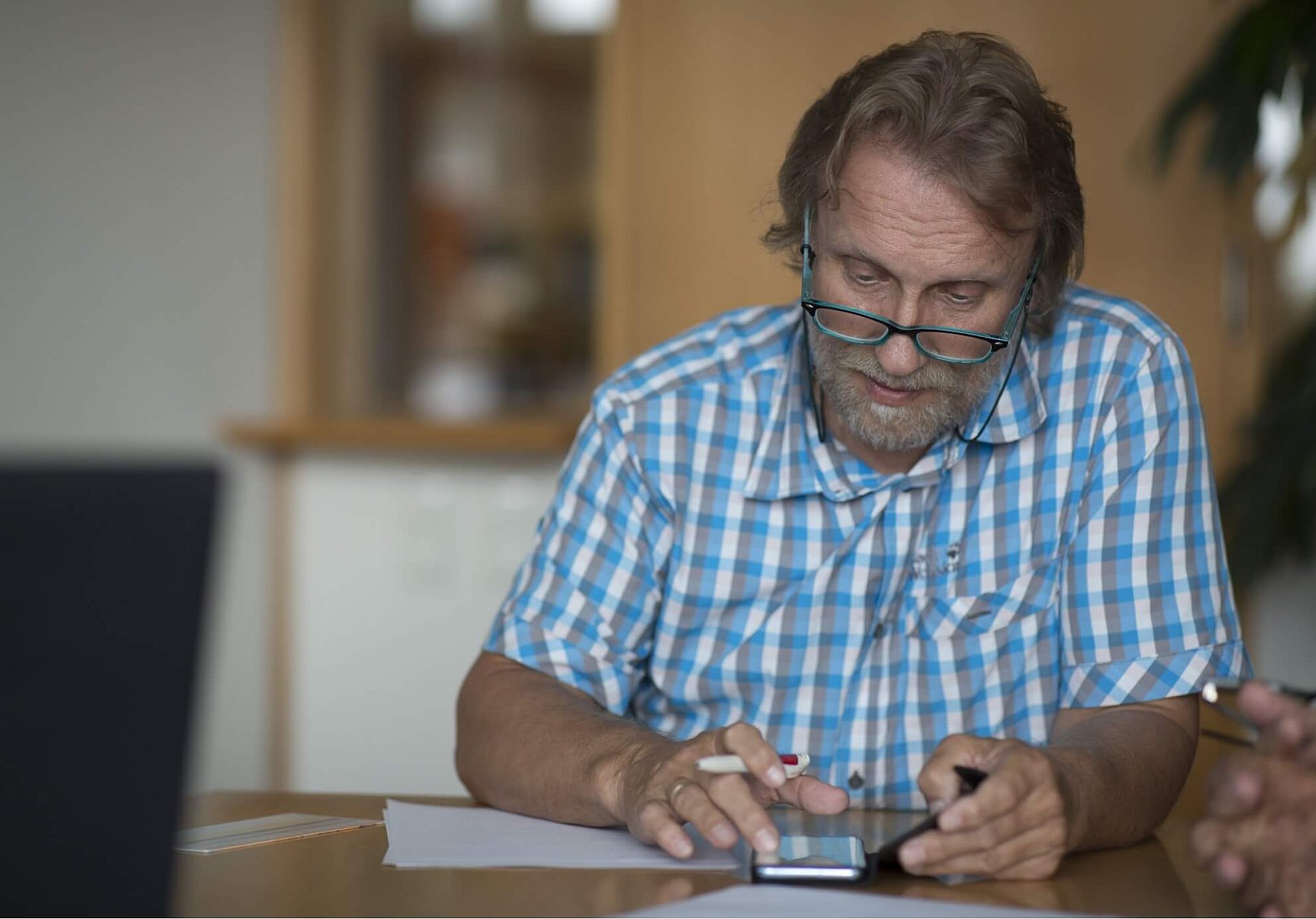  A middle-aged man with glasses and a blue chequered shirt is sitting at a table, looking down and using a smartphone. There are papers on the table in front of him. 