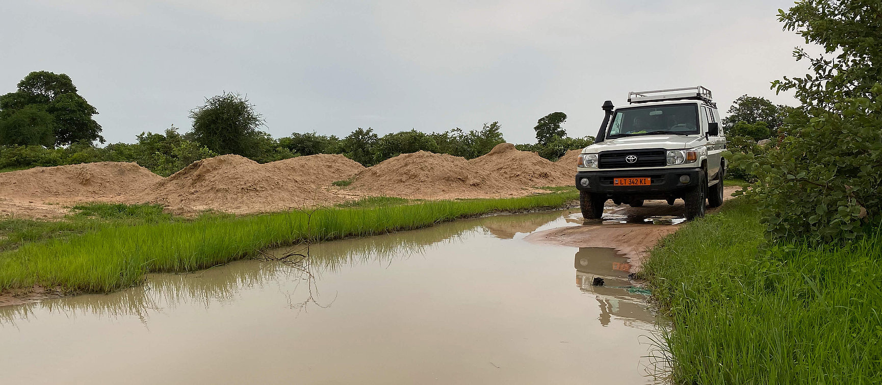 A white off-road vehicle next to a large puddle on a dirt road, with piles of sand and green vegetation in the background under a cloudy sky. 