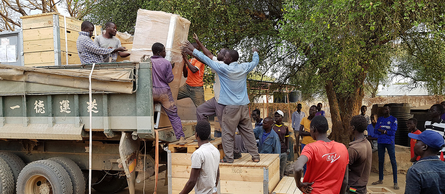 Several men unload large crates from a lorry while others watch and help outside, surrounded by trees and wooden crates. The scene has a communal feel. 