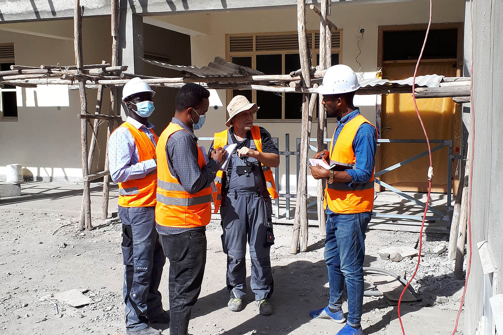 Four construction workers in orange-coloured safety waistcoats and helmets stand in a group and talk on a building site. Scaffolding and a half-finished building can be seen in the background. 