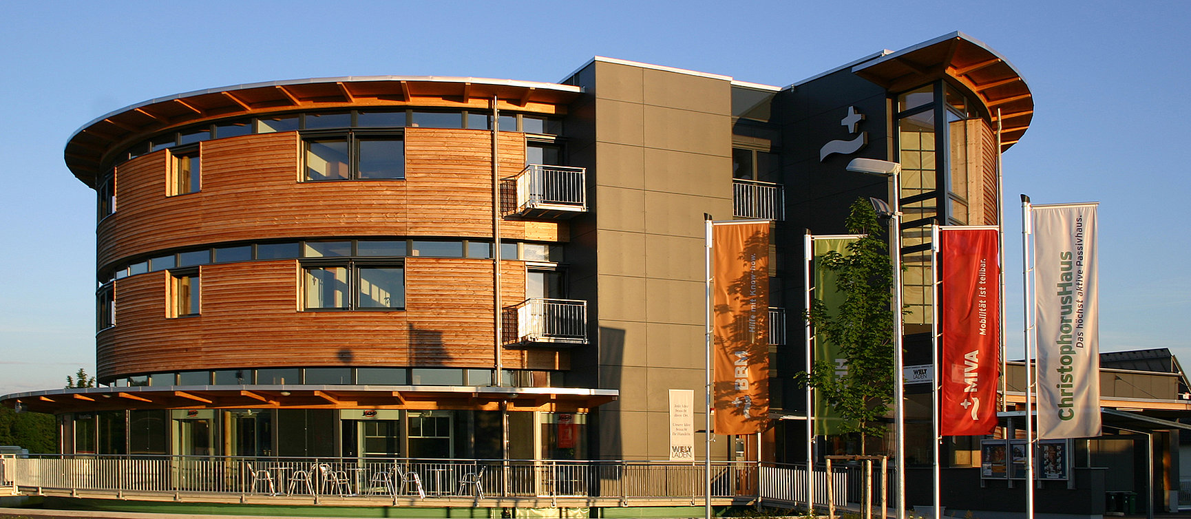 The ChristophorusHaus, a modern building, combines wood and grey panels in the façade. Large windows break through the front, while four colourful flags wave in front of it in the sunlight. 