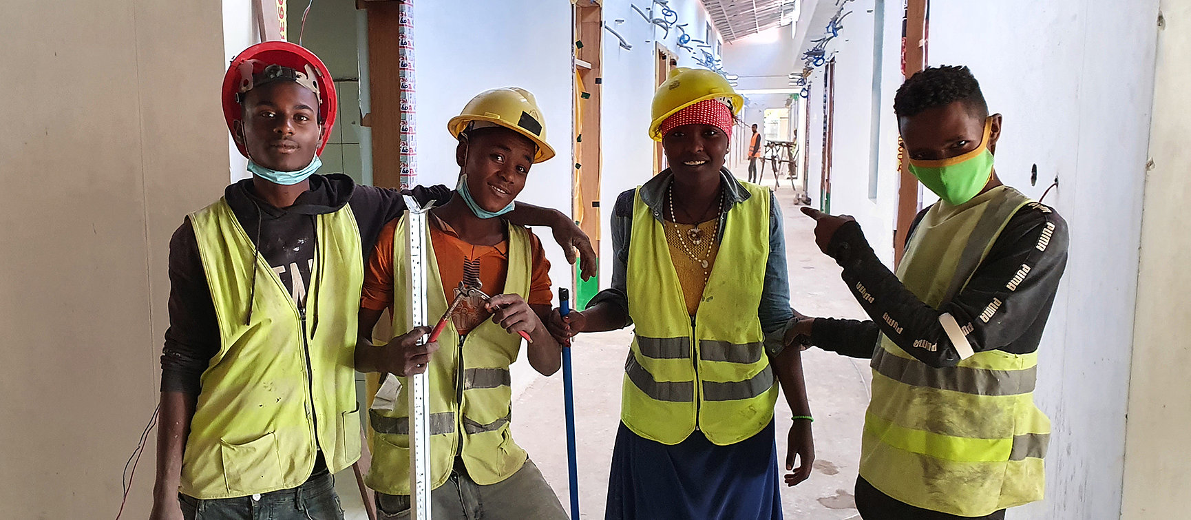 Four construction workers in yellow safety waistcoats and helmets stand together in the building, smiling and posing for the camera. Some are wearing face masks under their chins. Tools can be seen in their hands.