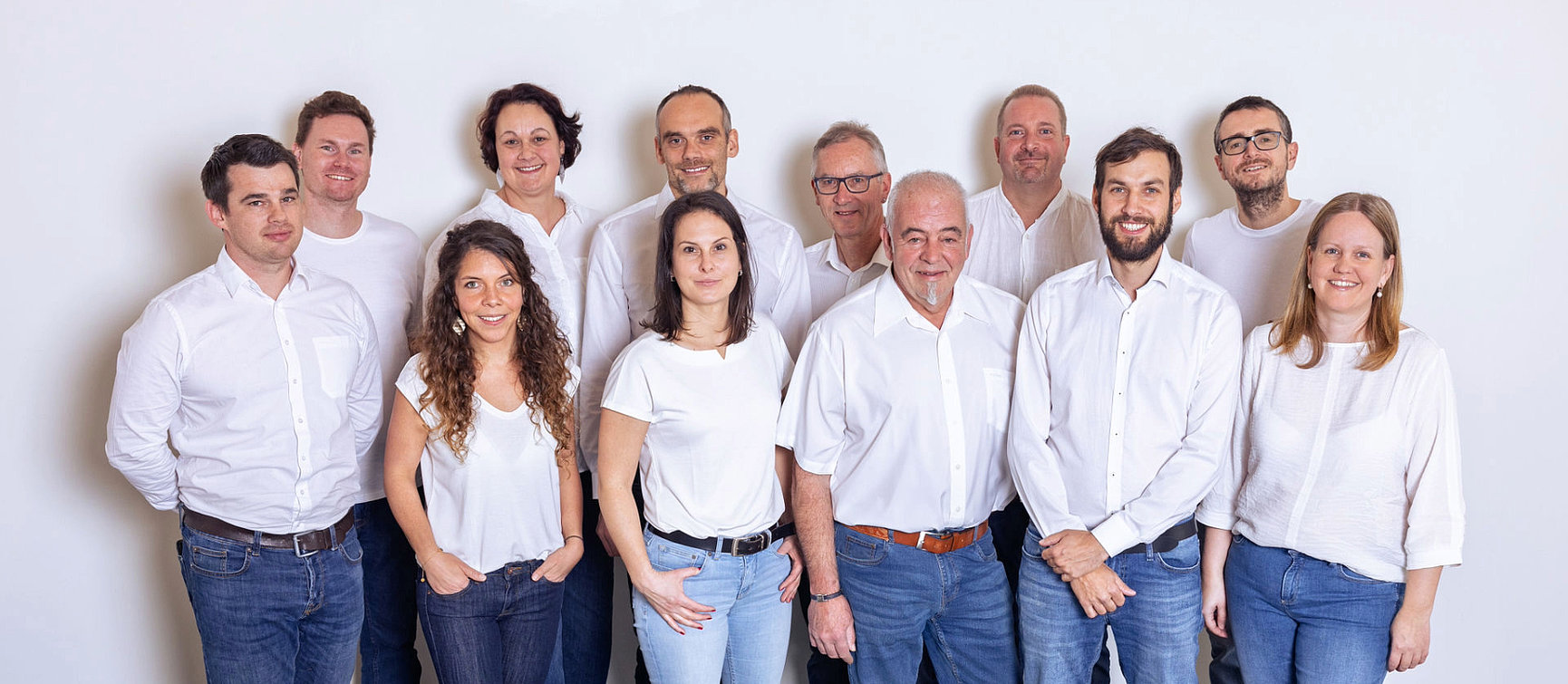 The BBM team, consisting of thirteen men and women, stands in front of a plain white background.They are all wearing white tops and jeans and smiling in a friendly manner. 