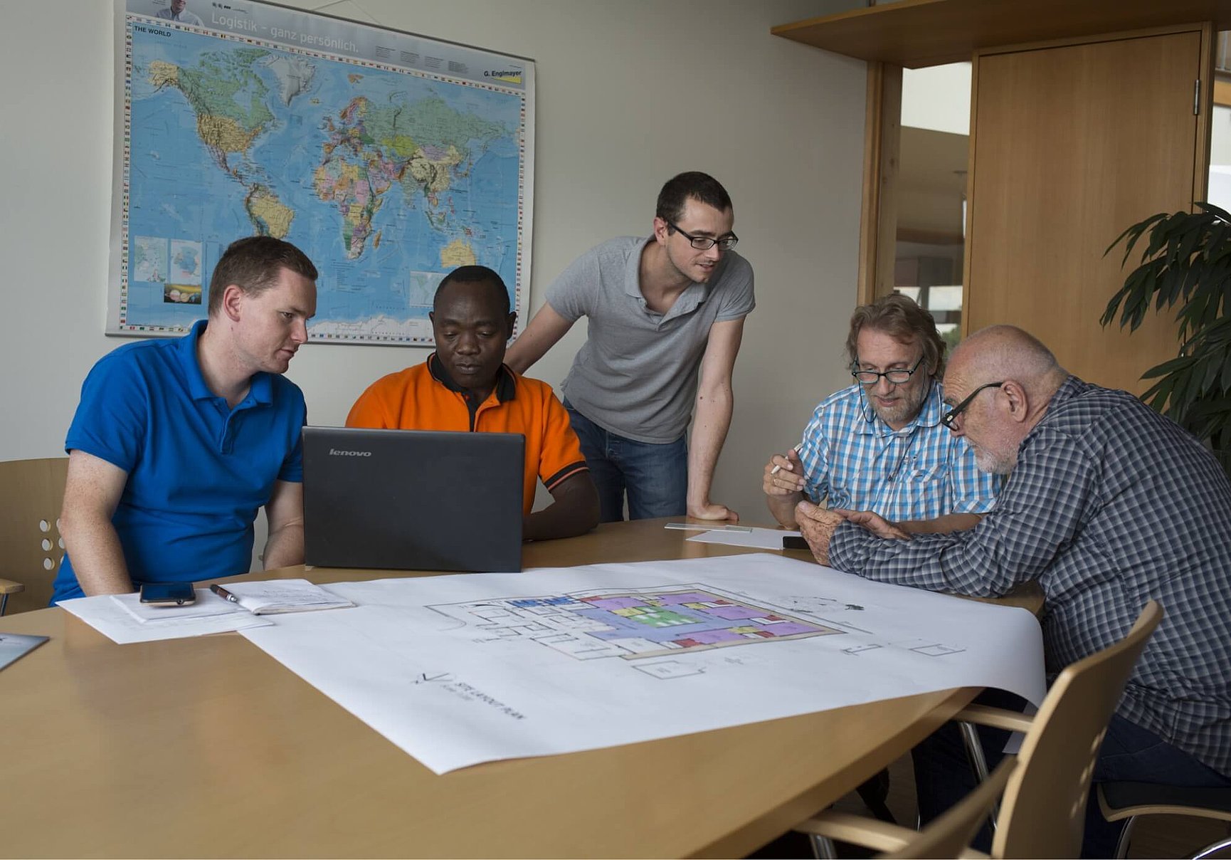 Five men sit around a table with architectural plans and a laptop, discussing. Behind them, a world map hangs on the wall in a bright, modern office.
