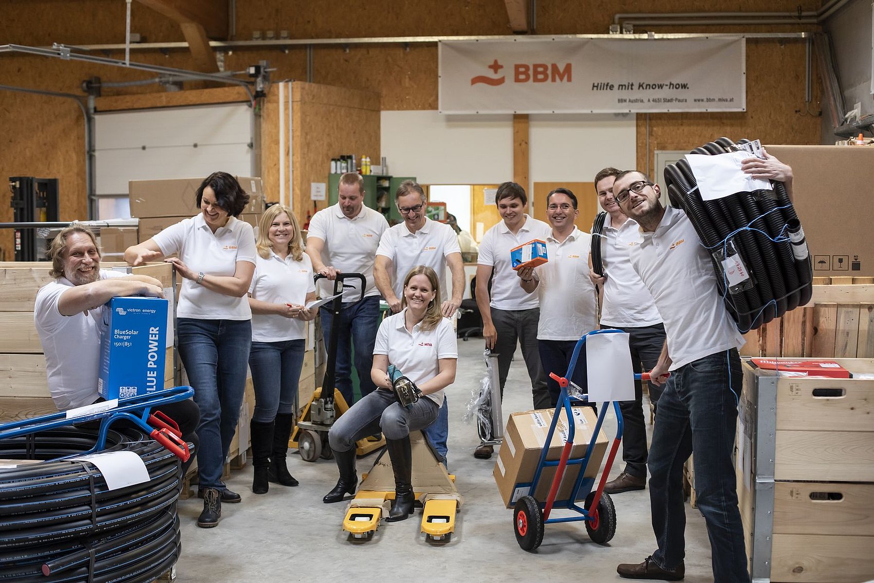 The BBM team poses smiling in a warehouse. The team members wear white shirts and hold tools or equipment in their hands. Some are standing, others are kneeling or working with trolleys. A banner with the BBM logo hangs on the wall in the background. 