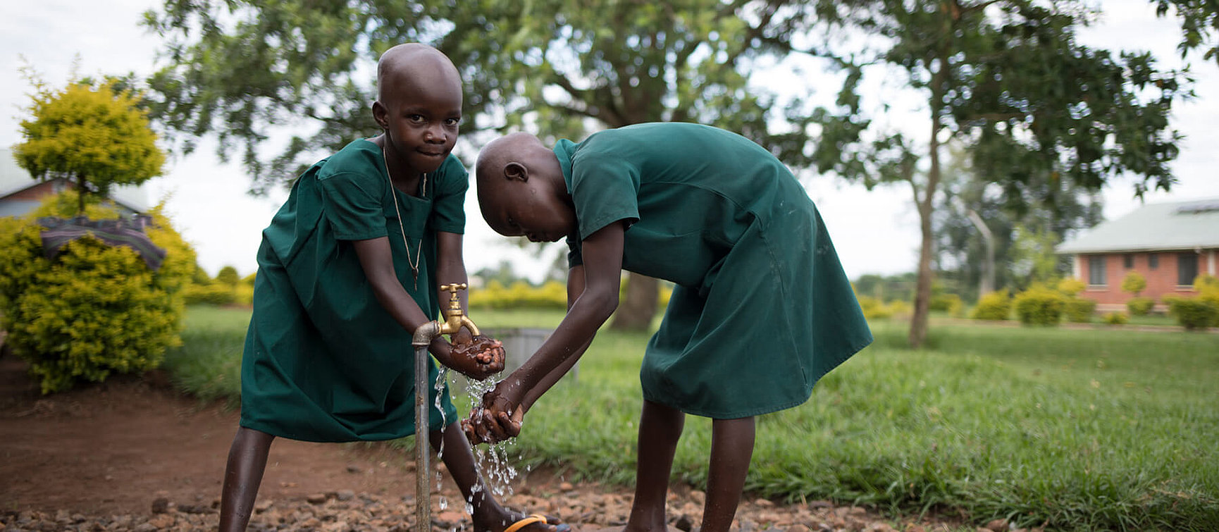 Two small children in green uniforms wash their hands together at an outdoor tap on a lawn. Trees and buildings can be seen in the background. 