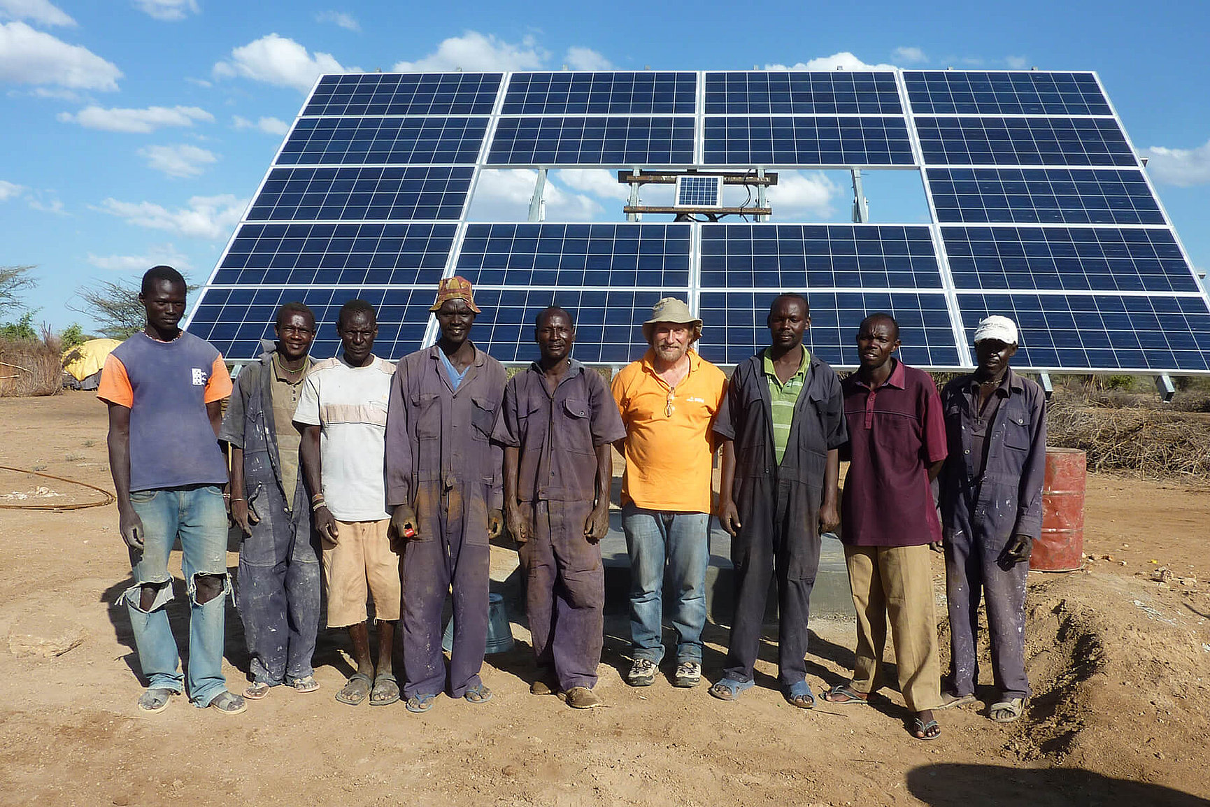 A group of nine men, some in work overalls and one in an orange shirt and hat, stand smiling in front of a large row of solar panels outside on a sunny day. 