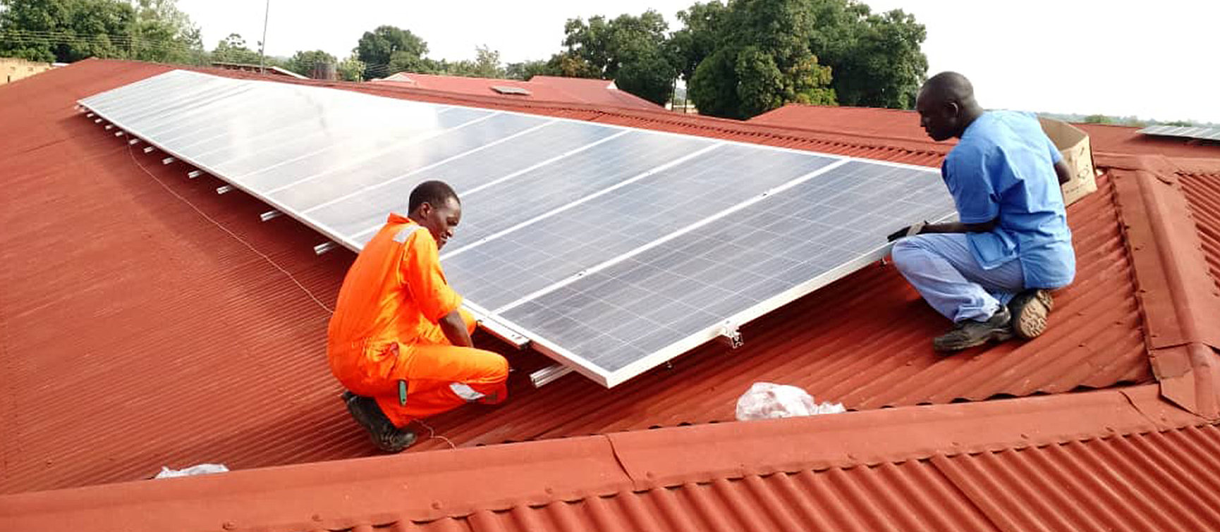 Two workers, one in an orange-coloured overall and the other in blue clothing, install solar modules on a red metal roof. Trees can be seen in the background.