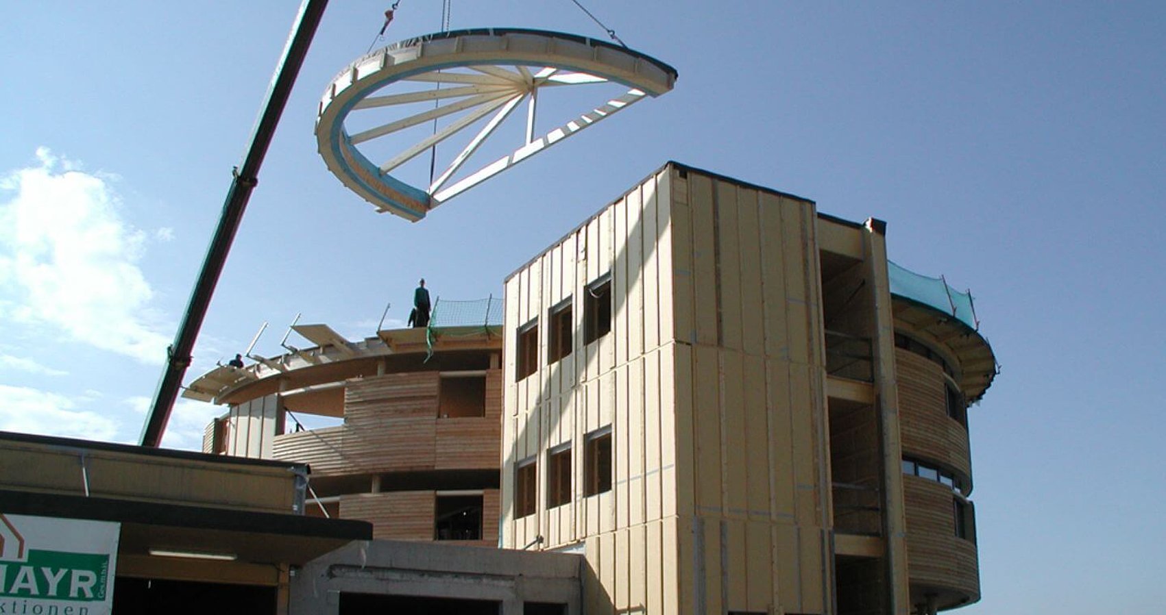 A crane lifts a large, curved roof section onto the roof of a partially completed multi-storey building. A worker stands on the roof, the clear blue sky can be seen in the background. 