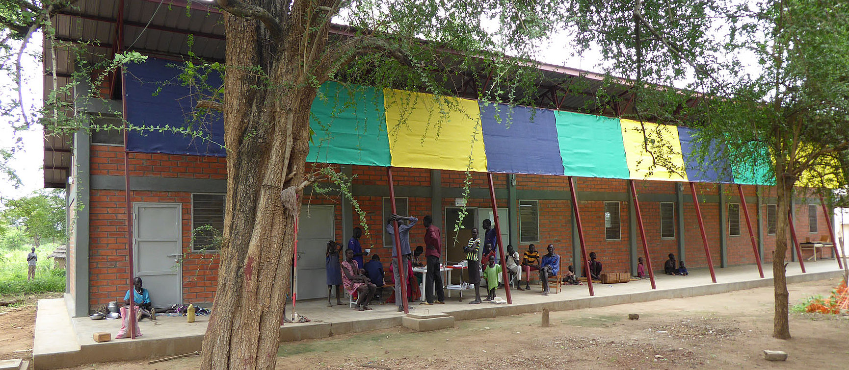 A group of people, including children, sit and stand on unpaved ground in front of a brick building with colourful panels and a metal roof shaded by trees.