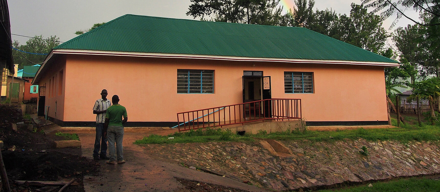 Two people stand on a path in front of a one-storey peach-coloured building with a green roof, barred windows and a red ramp leading to the entrance, surrounded by trees and greenery.