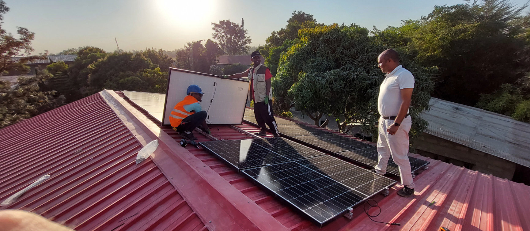 Three men are installing solar modules on a red metal roof surrounded by trees. One man is wearing an orange-coloured safety waistcoat and a helmet, the others are wearing casual clothes. 