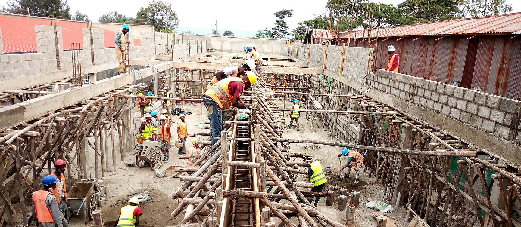 Construction workers wearing safety waistcoats and helmets erect a structure made of wooden scaffolding, concrete blocks and metal reinforcements on a building site. Some are standing on the ground, others on scaffolding.