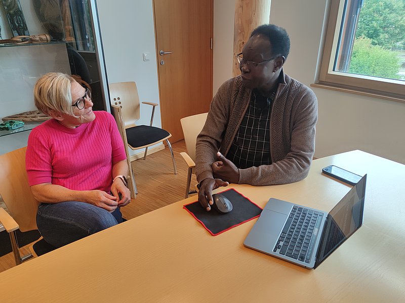 A woman in a pink jumper and a man with glasses are sitting at a table with a laptop and phone, discussing PROSUDAN projects in South Sudan in a bright office with wooden chairs and a window.