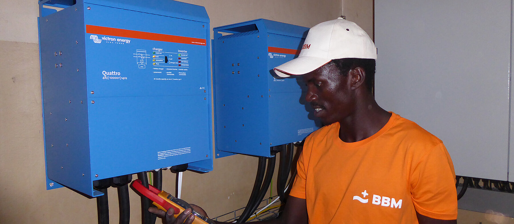 A man in an orange BBM shirt and a white cap uses a tool to check a blue Victron Energy Quattro inverter mounted on a wall 