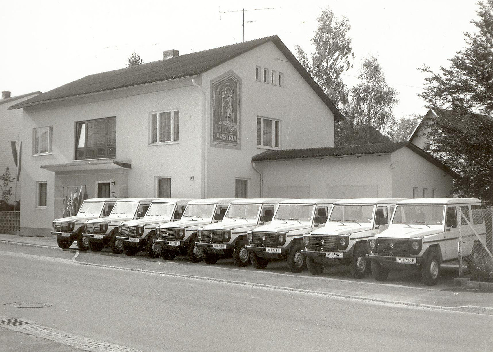 A row of eight vintage SUVs is parked in front of a two-storey building with a religious mural on the façade. Trees and bushes line the sides of the building. The picture is black and white. 
