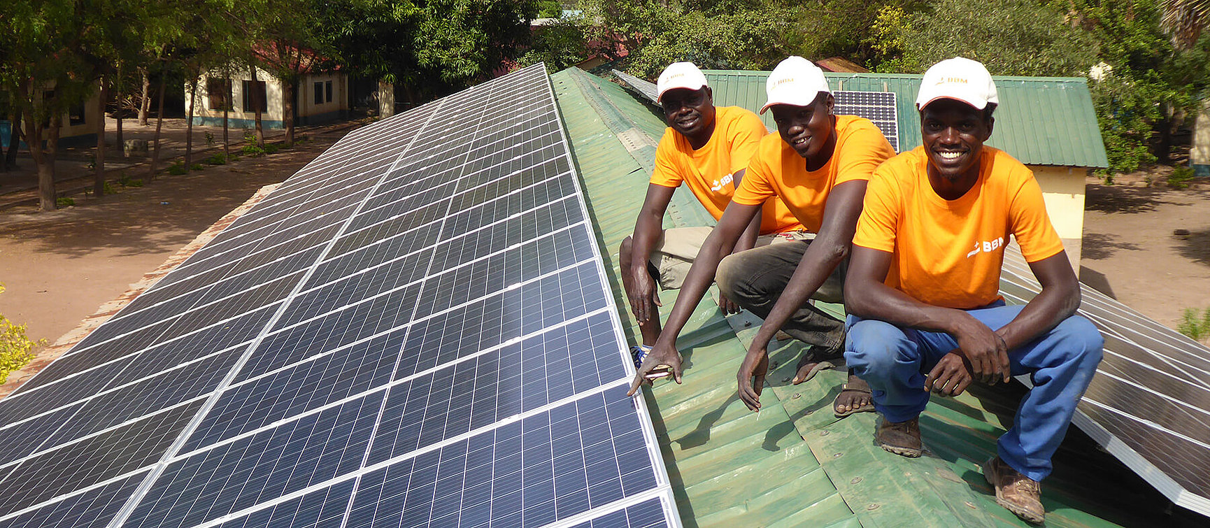 Three men in orange shirts and white caps smile as they install or inspect solar modules on a green metal roof with trees and buildings in the background. 