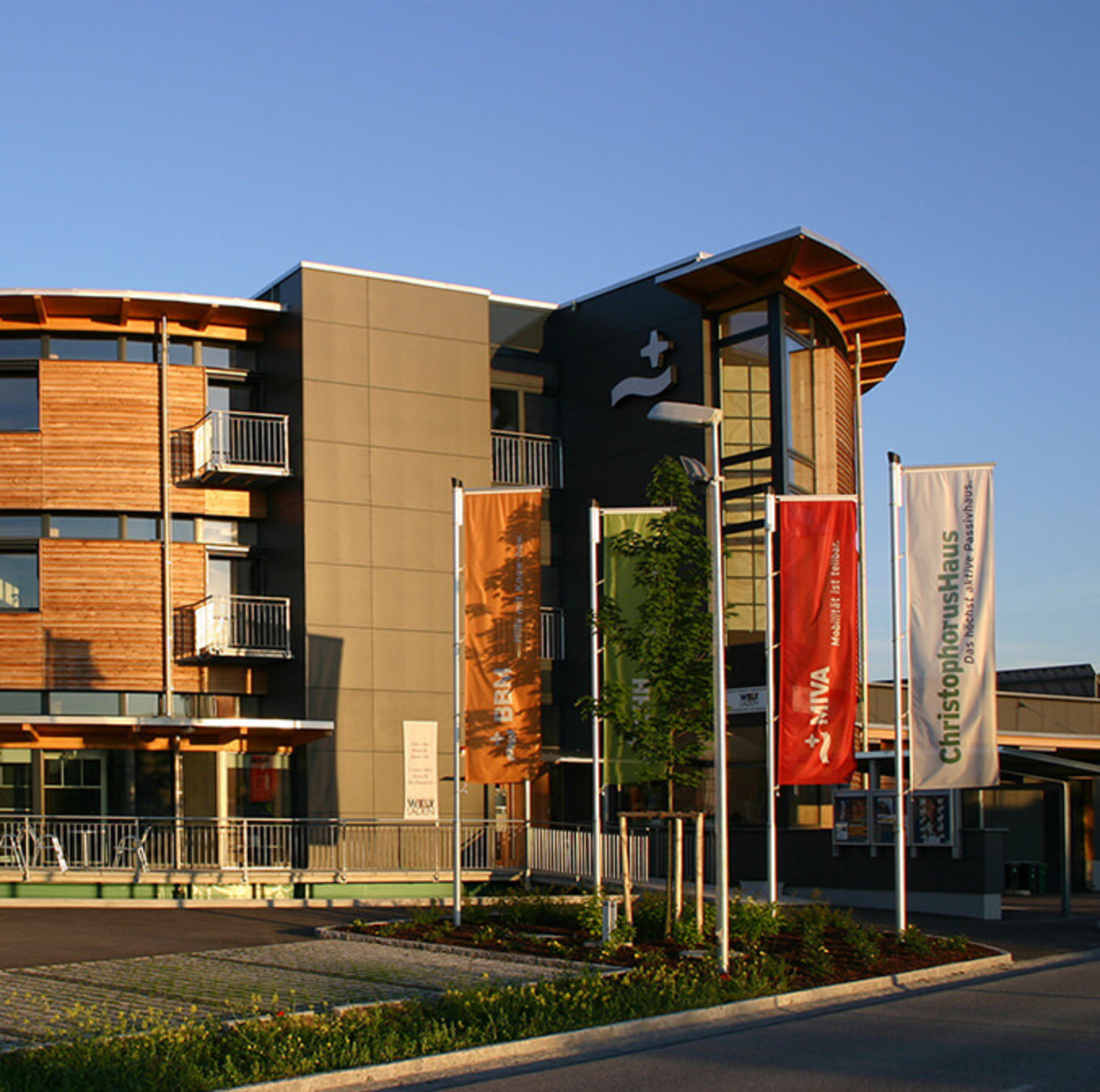 The ChristophorusHaus, a modern building, combines wood and grey panels in the façade. Large windows break through the front, while four colourful flags wave in front of it in the sunlight. 