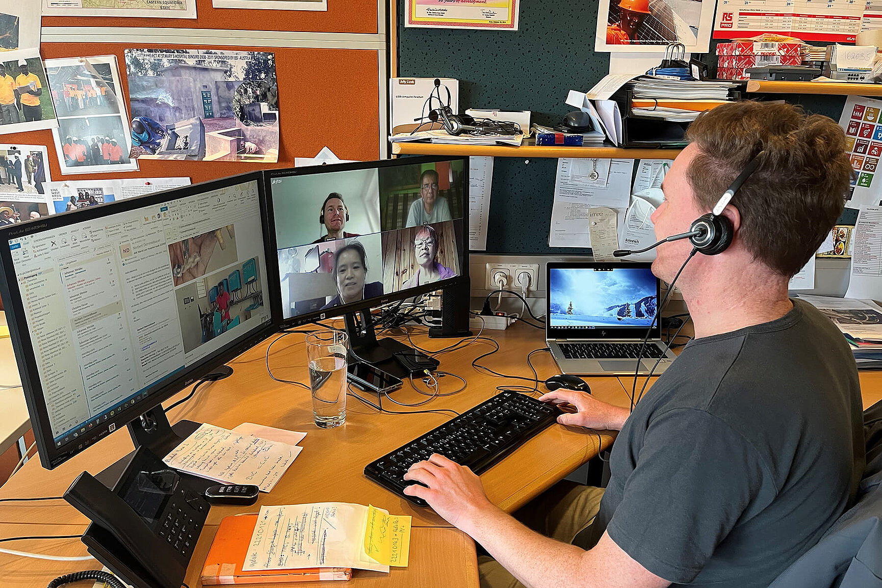 A man with a headset is sitting at a desk with two monitors. A video call with five people is running on one of them. Papers, a glass of water and electronic devices lie on the desk. Pinboards and photos hang on the wall behind him. 