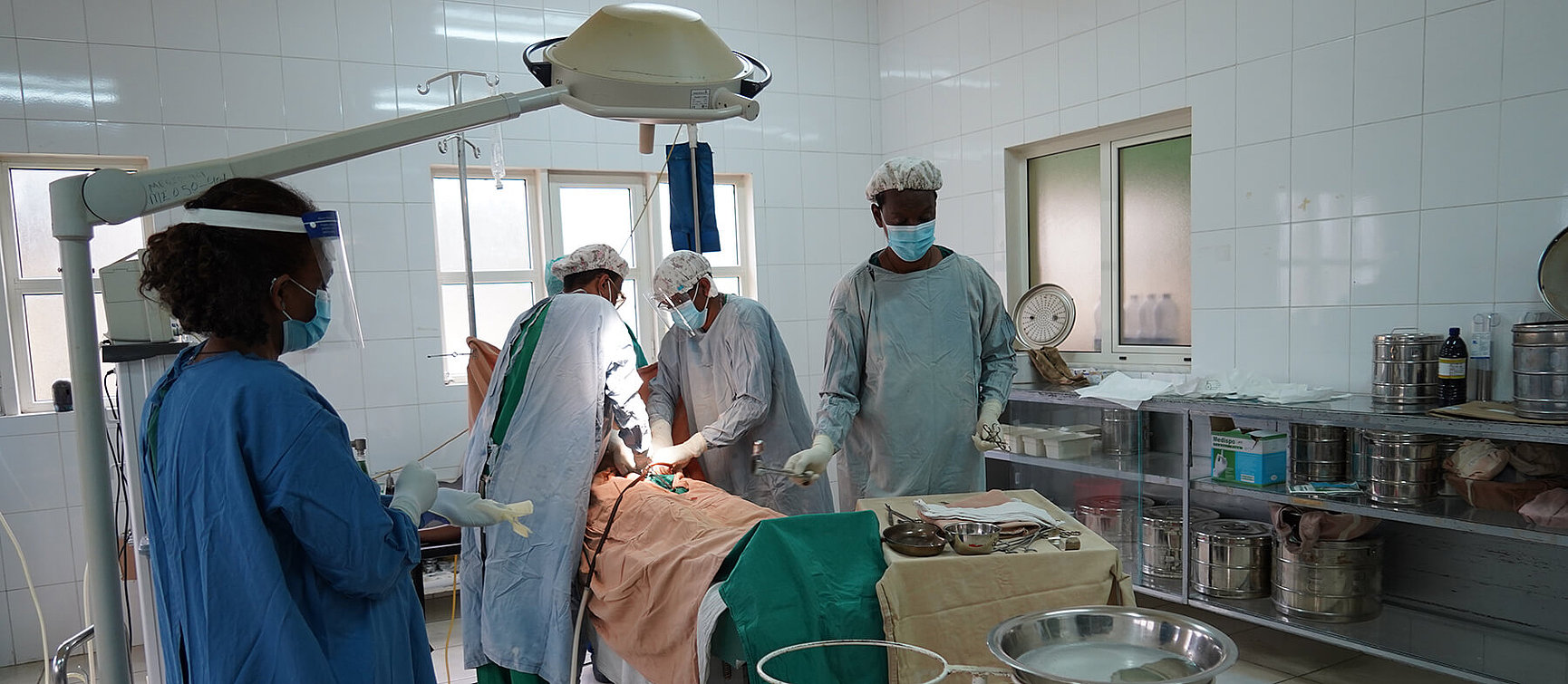 Several medical professionals in surgical clothing and wearing face masks operate on a patient in a well-lit operating theatre. Medical equipment and instruments are located nearby. 