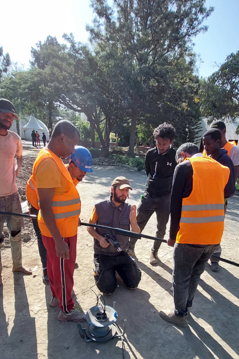 Three men are installing solar modules on a red metal roof surrounded by trees. One man is wearing an orange-coloured safety waistcoat and a helmet, the others are wearing casual clothes.