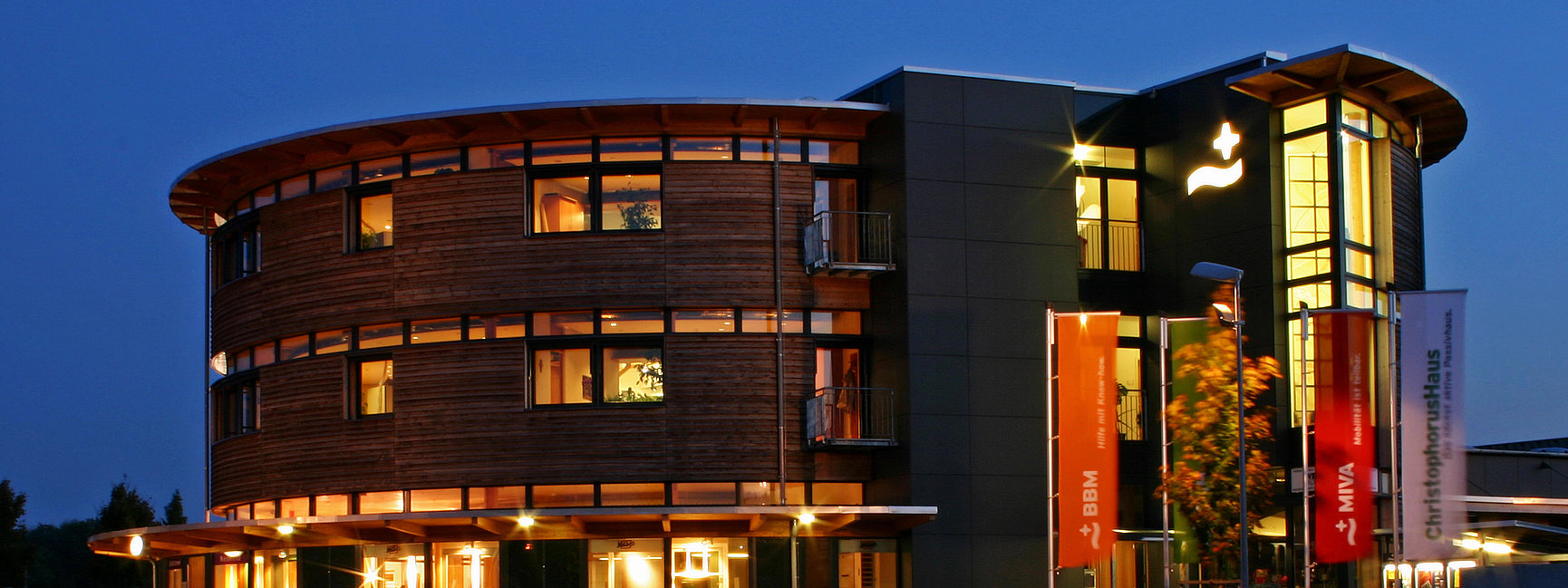 ChristophorusHaus, a building with curved wood panelling and dark walls, illuminated windows and orange flags in front of it, photographed at dusk against a clear evening sky.