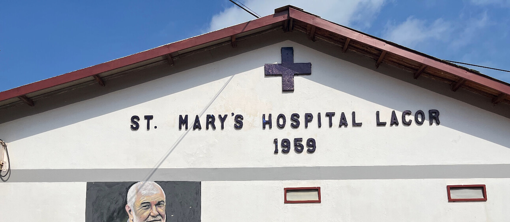 The picture shows the front of a building with the sign ‘St Mary's Hospital Lacor 1959’ and a cross above the text, under a sloping roof against a blue sky.