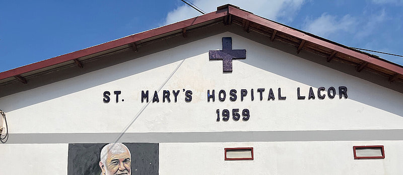 The front of a building with the sign ‘St Mary's Hospital Lacor 1959’ and a cross above the text, under a sloping roof against a blue sky.