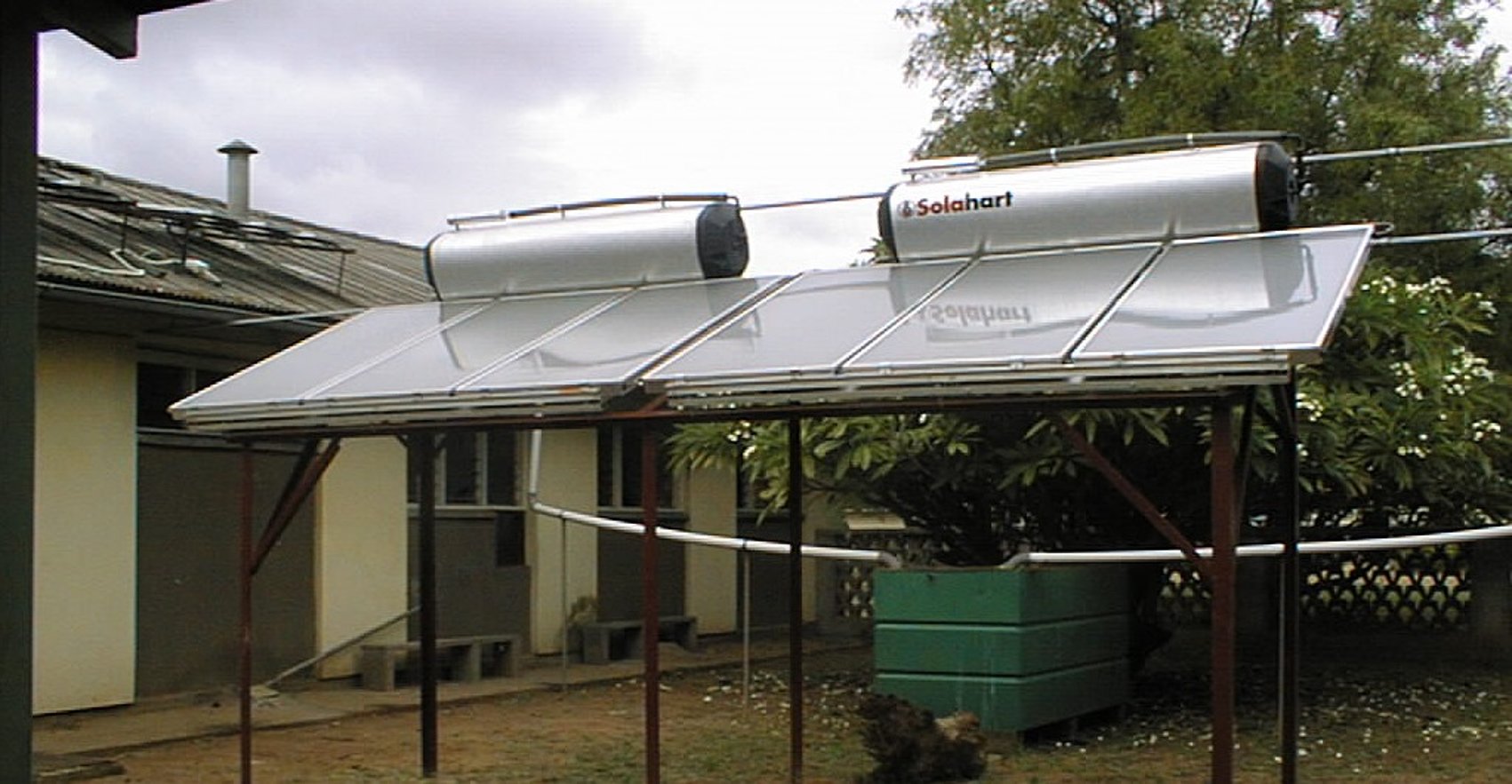 Two solar water heaters on the roof labelled ‘Solahart’ are mounted above a metal frame with large, tilted solar modules and are located in the courtyard of a house with trees. 