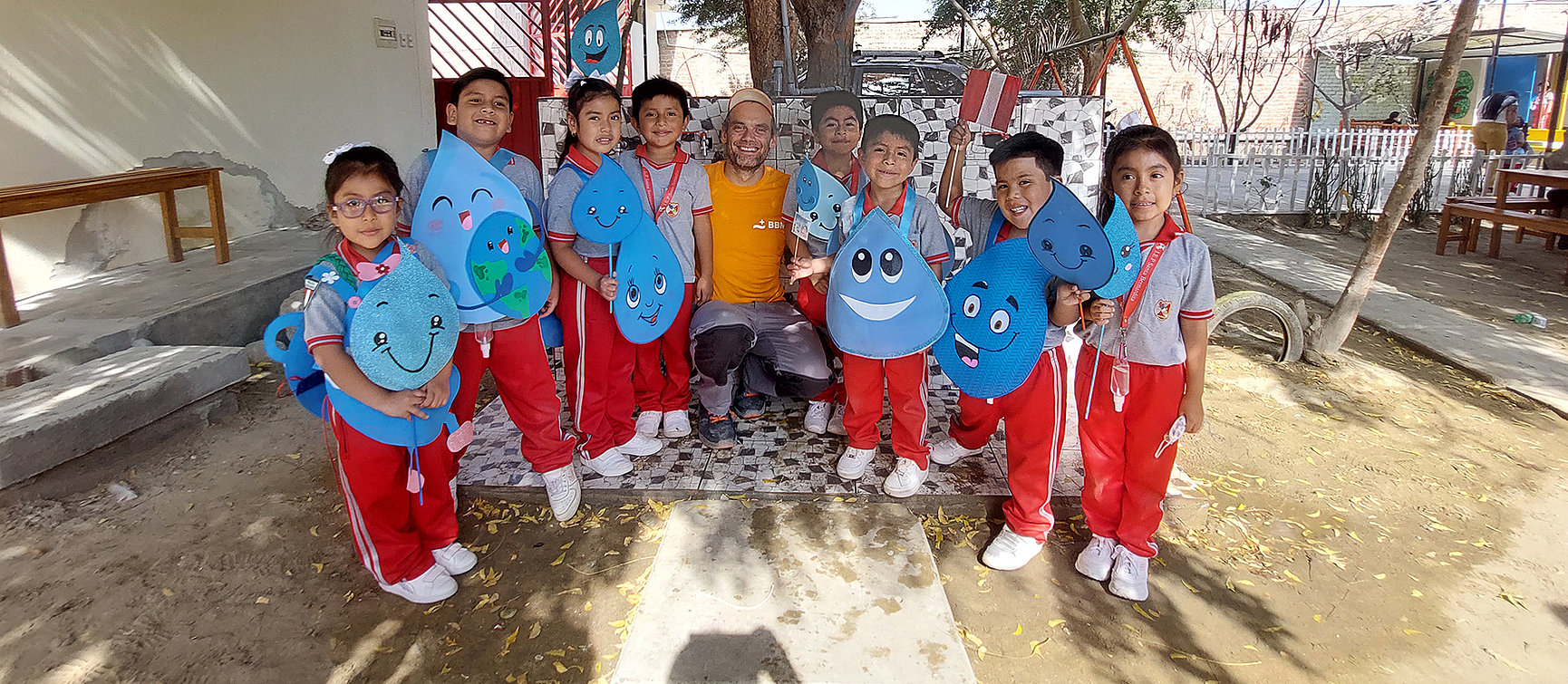 A group of young children in red and white uniforms pose outside with two adults. The children are holding large, decorated blue water drop cut-outs with smiling faces. Trees, benches and play equipment can be seen in the background.